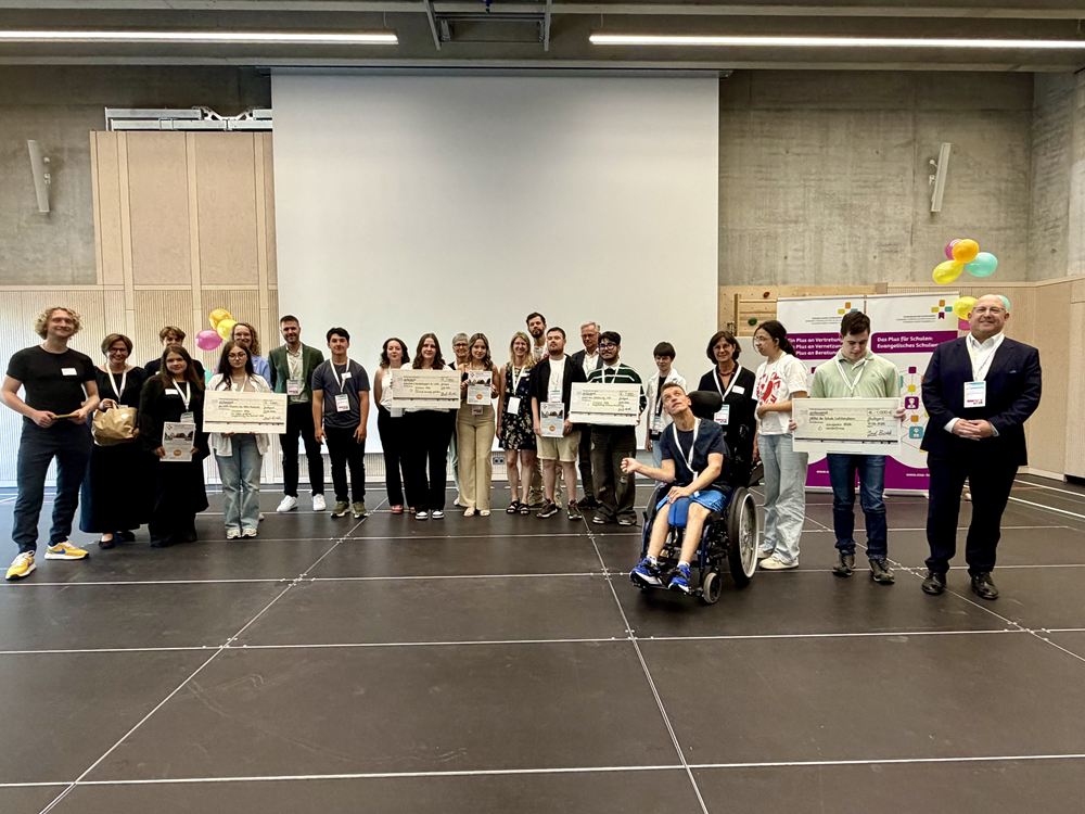 Auf der Bühne steht eine größere Gruppe von Frauen und Männern nebeneinander in einer Reihe und blickt in die Kamera. Mehrere Personen halten große Plakate oder Urkunden in den Händen. In der Mitte vorne befindet sich eine Person im Rollstuhl. Der Hintergrund besteht aus einer großen Leinwand in der Mitte und hellgrauen Wänden. Rechts und links im Hintergrund hängen bunte Luftballons an einer Holzwand, daneben stehen zwei Roll-ups. Der Boden der Bühne ist dunkel.