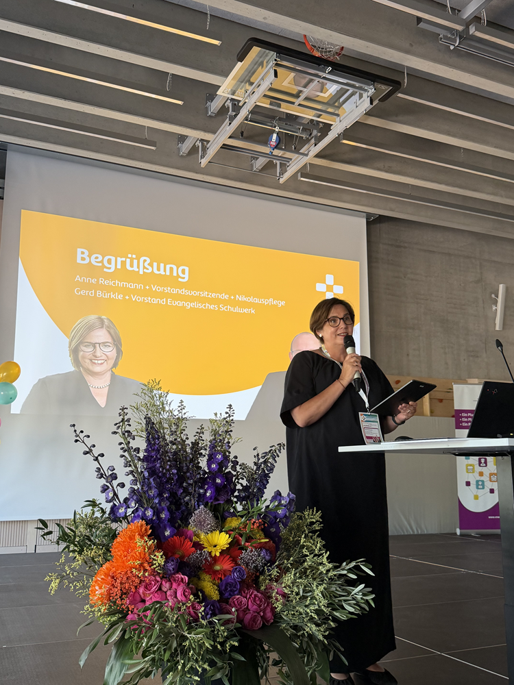 Auf dem Foto ist eine Frau auf einer Bühne an einem Rednerpult mit Mikrofon zu sehen, daneben ein großer bunter Blumenstrauß im Vordergrund. Hinter der Person hängt eine Leinwand mit einer gelb-weißen Präsentationsfolie, auf der links ein Porträtfoto derselben Person und darüber Text steht. Die Schrift in weiß lautet: „Begrüßung Anne Reichmann + Vorstandsvorsitzende + Nikolauspflege Gerd Bürkle + Vorstand Evangelisches Schulwerk“.